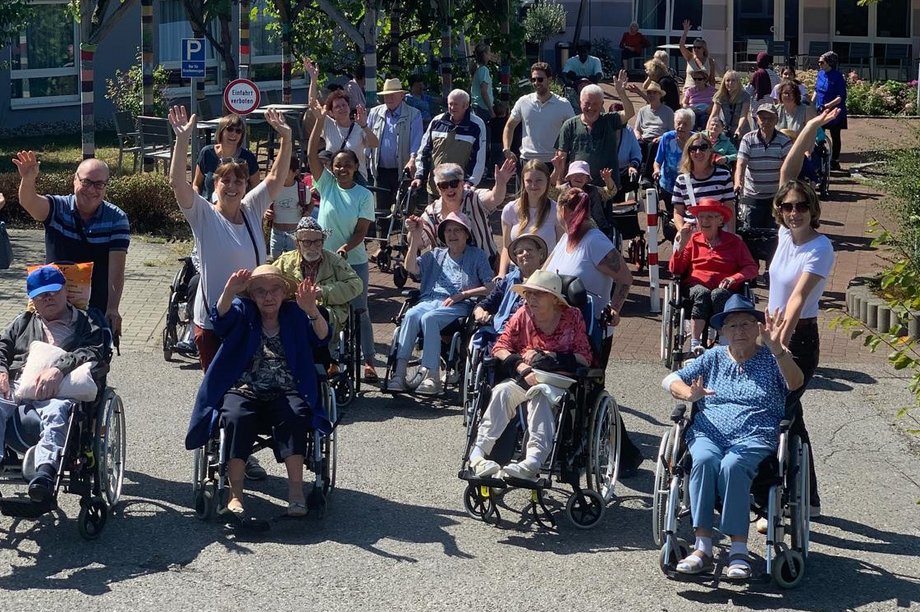 Bewohnerinnen und Bewohner eines Pflegeheims unternehmen gemeinsam mit Mitarbeitenden einen Ausflug im Freien und winken in die Kamera.