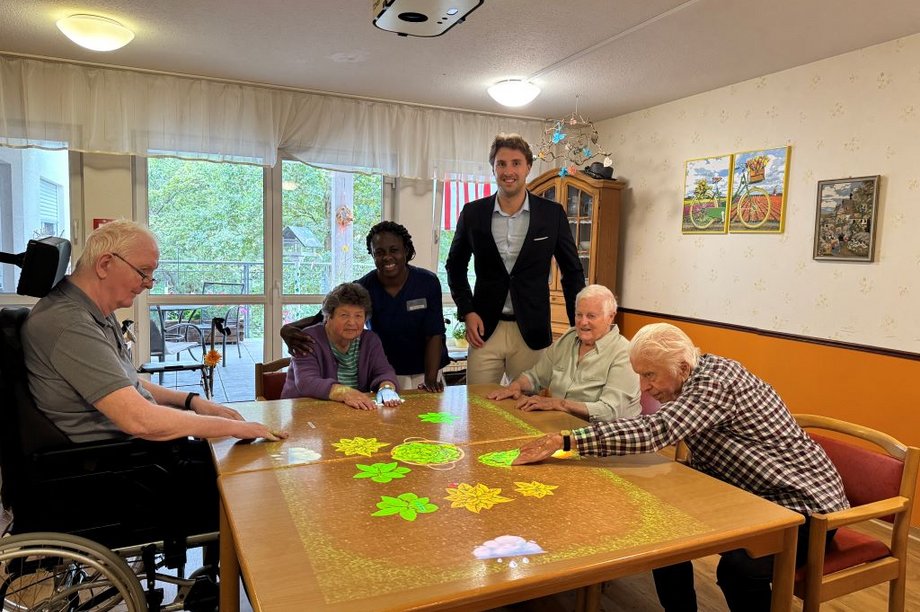 Vier Bewohnerinnen und Bewohner des AWO-Seniorenzentrums Rheinaue sitzen an der neuen Tovertafel. Daneben stehen eine Pflegekraft sowie Einrichtungsleiter Marvin Winnes.