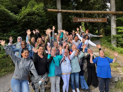 Mitarbeitdende des AWO-Seniorenzentrums Rheinaue stehen mit ausgestreckten Händen unter einem Schild auf dem Baumwipfelpfad steht.