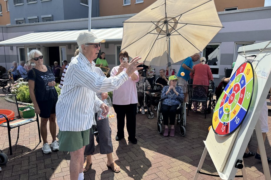 mehrere Seniorinnen und Senioren sitzen vor Seniorenzentrum und ein Mann wirft ein Ball auf eine Klettzielscheibe.