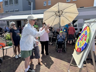 mehrere Seniorinnen und Senioren sitzen vor Seniorenzentrum und ein Mann wirft ein Ball auf eine Klettzielscheibe.